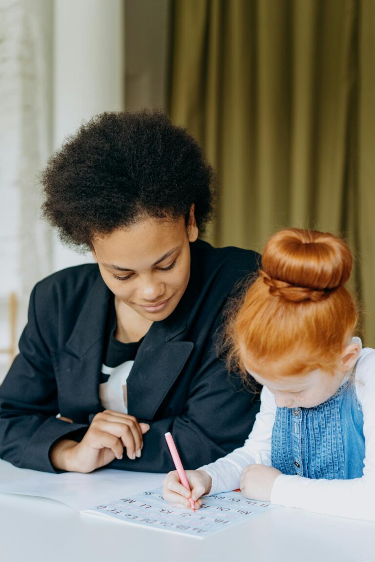 A teacher assists a student with her homework at a desk indoors, promoting learning and education.